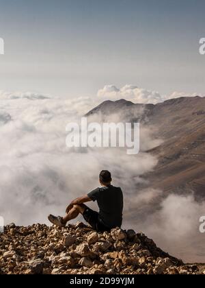 uomo seduto su una cima che si affaccia su una vista panoramica una catena montuosa e le nuvole che coprono Foto Stock