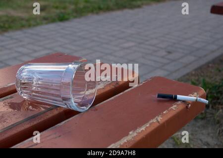 Una sigaretta fumatrice gettata su una panca marrone e un vuotato vetro su uno sfondo di erba verde e un marciapiede grigio Foto Stock