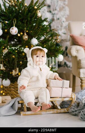 Carino divertente un bambino di un anno in un morbido jumpsuit con le orecchie sul cappuccio si siede su una slitta di legno giocattolo a. L'albero di Natale Foto Stock