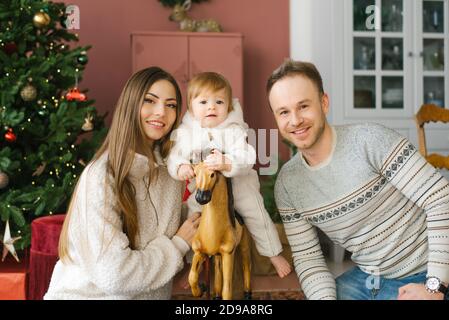 Un bambino piccolo di un anno è seduto su un cavallo di legno vicino all'albero di Natale. I suoi genitori sono seduti accanto a lui. Felice ritratto di famiglia Foto Stock