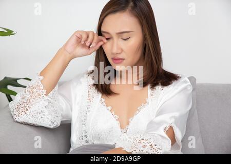 Le donne asiatiche negli indumenti da notte in raso bianco stanno graffiando le loro palpebre a causa dell'allergia in una stanza. La femmina ha un occhio prurito. Il concetto di allergia Foto Stock