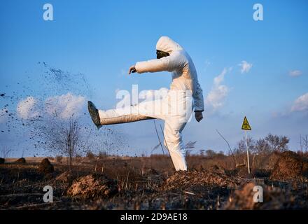 Vista laterale orizzontale istantanea di uno scienziato deluso, indossando un vestito protettivo bianco, maschera a gas e guanti, calci il terreno su campo bruciato contro il cielo blu Foto Stock