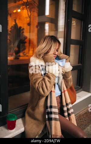 Bella ragazza bionda la sensazione di freddo, cercando di proteggere se stessa dal congelamento della città sul giorno d'inverno. Giovane donna caucasica attesa per amico sulla s Foto Stock