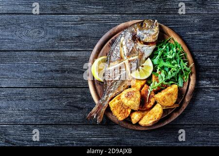 Pesce dorado arrosto intero con pelle su patata al forno in pangrattato servito su un tagliere di legno con limone cuneo e rucola fresca o selvatica Foto Stock