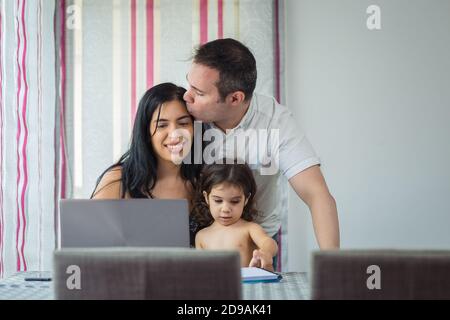 Madre ispanica e padre caucasico baciano felicemente con la loro bambina sul loro giro dipingendo sul notebook, con un portatile di fronte a loro, seduto Foto Stock