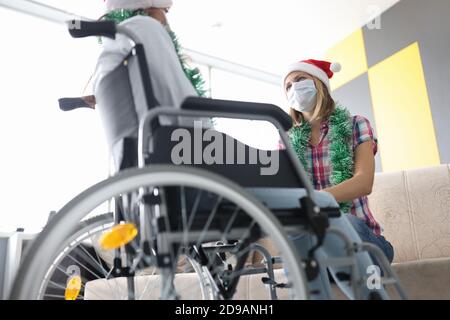 Donna siede in sedia a rotelle nel cappello di babbo natale di fronte alla donna in maschera medica protettiva Foto Stock