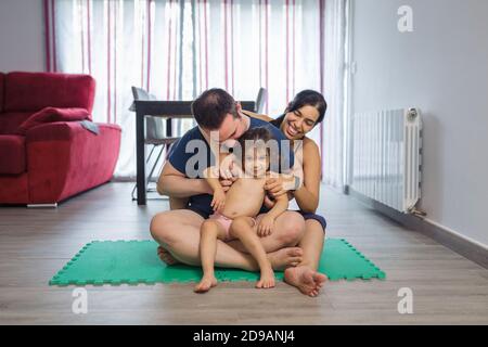 Il padre caucasico, la madre ispanica e la bambina sono seduti su un tappetino yoga, vestiti con abiti sportivi e abbracciati, foto ritratto nella sala da pranzo di Foto Stock