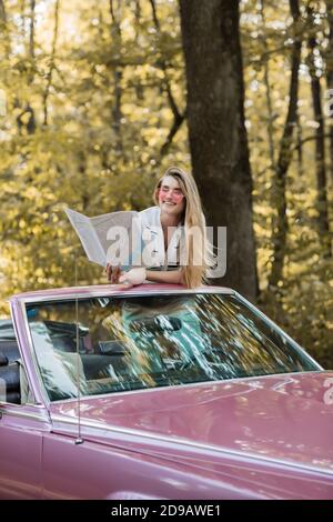 donna sorridente in occhiali da sole che guarda l'atlante stradale mentre si siede in cabriolet in foresta Foto Stock