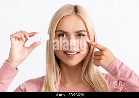 Donna sorridente che punta con il dito all'occhio mentre tiene la scatola con lenti a contatto isolate su bianco Foto Stock