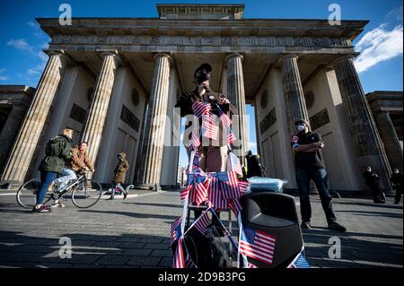 Berlino, Germania. 04Nov 2020. Un partecipante che indossa un protettore per bocca e naso parla in un raduno alla porta di Brandeburgo sotto il motto "transizione pacifica della Presidenza e di un democratico degli Stati Uniti" accanto a una scala a cui sono attaccate piccole bandiere statunitensi. Più di 200 milioni di americani sono stati chiamati ad eleggere un nuovo presidente e i membri della Camera dei rappresentanti. Credit: Fabian Sommer/dpa/Alamy Live News Foto Stock