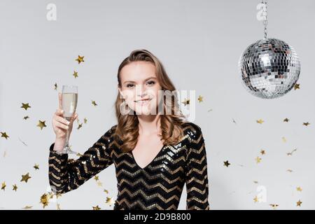 giovane donna allegra che tiene un bicchiere di champagne vicino alla palla da discoteca e i confetti cadenti sul grigio Foto Stock