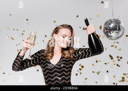 giovane donna allegra con bicchiere e bottiglia di champagne vicino palla da disco e confetti in caduta su grigio Foto Stock