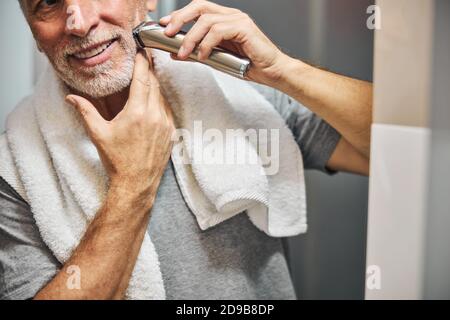 Uomo anziano sorride mentre si rade la barba con un dispositivo elettrico Foto Stock