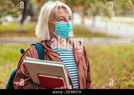Ritratto autunnale esterno di donna bionda che indossa maschera medica protettiva, vestiti casual, occhiali e uno zaino che tiene un libro di esercizi, notebook a piedi in strada Foto Stock