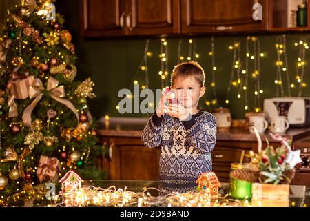 Un ragazzo si siede a un tavolo in una cucina di Natale decorata. Tenendo un giocattolo per decorare un albero di Natale. Accoglienti serate invernali a casa. Concetto di Cristo Foto Stock