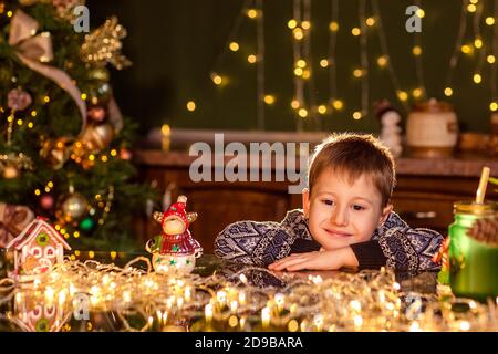 Un ragazzo si siede a un tavolo in una cucina di Natale decorata. Tenendo un giocattolo per decorare un albero di Natale. Accoglienti serate invernali a casa. Concetto di Cristo Foto Stock