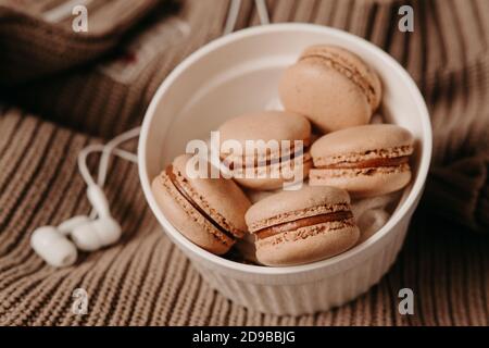 Cuffie bianche e macaroni al caramello su un maglione. Accogliente fotografia autunnale. Foto di cibo in tonalità marroni. Playlist invernale Foto Stock