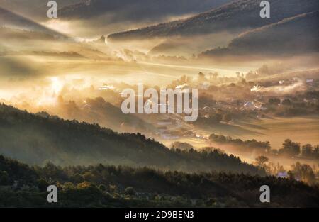 Vista aerea del villaggio in nebbia con raggi solari dorati all'alba in autunno. Bellissimo paesaggio rurale con strada, edifici, alberi colorati nebbia. Slovacco Foto Stock