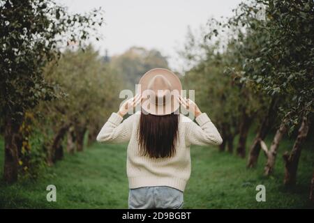 Vista posteriore di elegante donna dai capelli lunghi in cappello beige nel frutteto autunno. Foto Stock