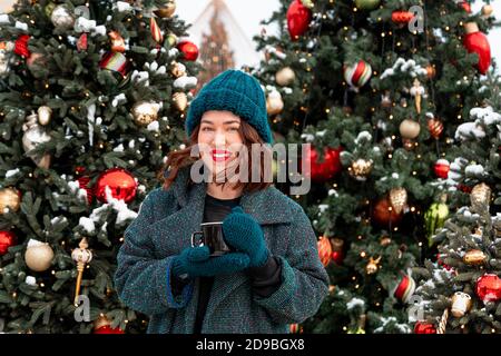 Bella Brunette Woman Holding Cup di cacao caldo o tè con albero di Natale su sfondo. 2021. Trascorrere le vacanze dell'anno NW con gioia e famiglia Foto Stock