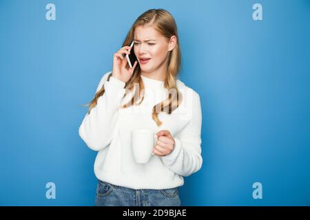 donna bionda confusa in maglione che parla con smartphone con la tazza isolato su sfondo blu Foto Stock