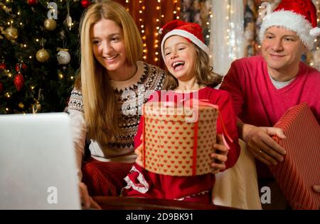 Una coppia felice con un bambino festeggia il Natale con i suoi amici in videochiamata con la webcam. La famiglia saluta i loro parenti alla vigilia di Natale Foto Stock