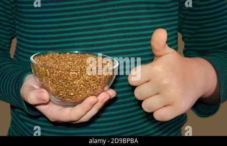 Il bambino tiene una lastra di vetro con grano saraceno nella sua mano destra, e mostra un simile con la sua mano sinistra. Primo piano. Dettagli. Foto Stock