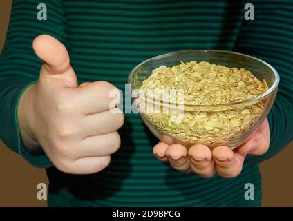 Il bambino tiene un piatto di vetro con farina d'avena nella mano sinistra, e mostra come con la mano destra. Primo piano. Dettagli. Foto Stock