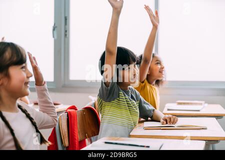 Il ragazzo asiatico di età elementare ha alzato le mani in su in Q ed IN UNA classe. Gruppo diversificato di alunni in età elementare nella scuola di costruzione dell'istruzione. Concetto di volontariato e partecipazione in classe. Foto Stock