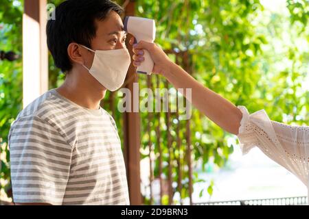 Primo piano mano utilizzando un termometro digitale a infrarossi medico su un giovane uomo in maschera facciale per controllare e monitorare la temperatura corporea. Nuova vita normale per lo screening dei visitatori per prevenire la diffusione di Coronavirus COVID-19 Foto Stock