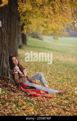 Bella donna in maglione, jeans e stivali seduta sotto l'albero d'acero con tazza di caffè o tè nel parco autunnale. Foto Stock