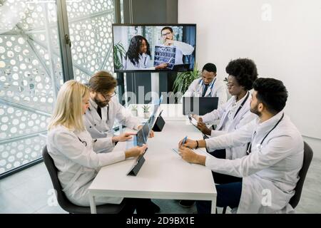 Un team di cinque medici multietnici diversi che si riuniscono in una videoconferenza in ospedale, chiacchierando con i due giovani colleghi africani e caucasici Foto Stock