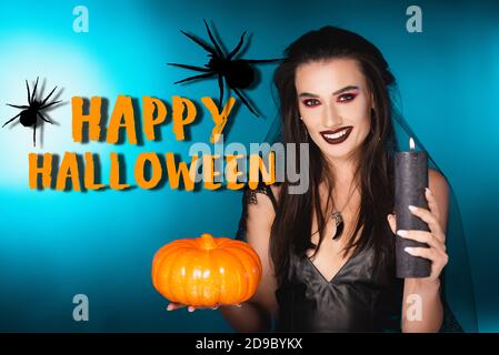 donna gioiosa con trucco nero e velo che tiene la candela bruciante e zucca vicino halloween felice scritta su blu Foto Stock