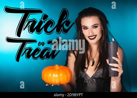 donna gioiosa con trucco nero e velo che tiene la candela bruciante e zucca vicino trucco o trattare la scritta blu Foto Stock