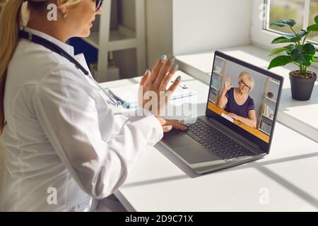 Dottore che sventola la mano sullo schermo del computer portatile dicendo ciao al senior donna durante la consultazione online Foto Stock