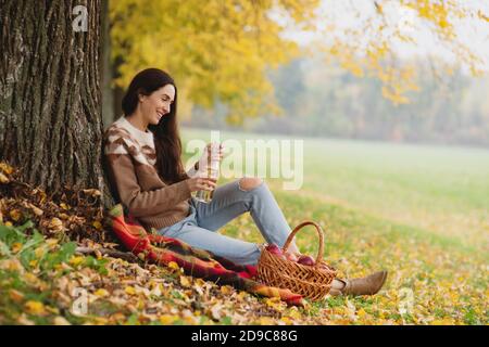 Ritratto di bella giovane donna seduta vicino all'albero nel parco autunnale e bevendo sidro da una bottiglia. Foto Stock