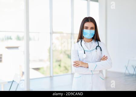 Donna medico indossando maschera medica con braccia incrociate in background ospedale. Spazio di copia. Medicina, professione e concetto di assistenza sanitaria Foto Stock
