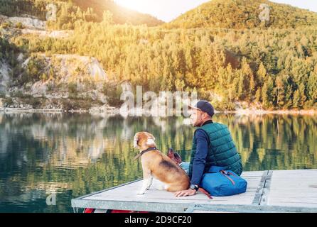 L'uomo proprietario del cane e il suo amico cane beagle sono seduti sul molo di legno sul lago di montagna e godere il paesaggio durante la loro passeggiata nel aut Foto Stock