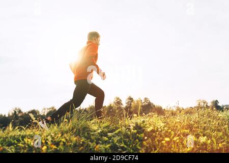 Un uomo vestito con camicia a manica lunga rossa, pantaloni da corsa neri e scarpe che corrono per la strada con uno sfondo cielo soleggiato Foto Stock