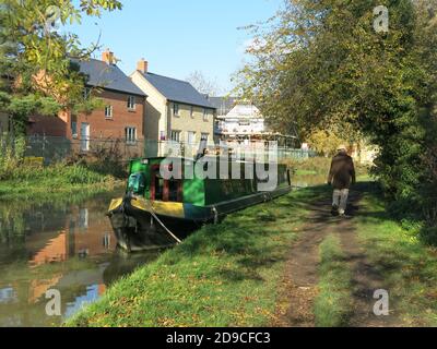 Un uomo cammina lungo l'alzaia passando un narrowboat che è ormeggiato di fronte alle nuove case che sono state costruite nel villaggio di Cosgrove, Northants. Foto Stock