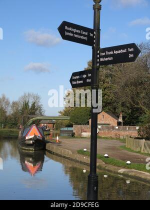 Vista del grazioso villaggio sul canale di Cosgrove sulla Grand Union, con una narrowboat ormeggiata sul sentiero e un cartello per i turisti. Foto Stock