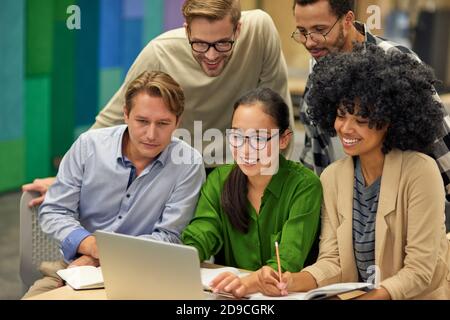 Gruppo di felici uomini d'affari multirazziali seduti alla scrivania nel moderno spazio di lavoro, guardando lo schermo del computer portatile e sorridendo, lavorando insieme. Concetto di collaborazione e lavoro di squadra Foto Stock