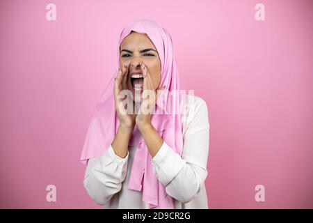 Giovane bella donna araba che indossa hijab islamico su rosa isolato gridando e gridando forte a fianco con le mani sopra bocca Foto Stock