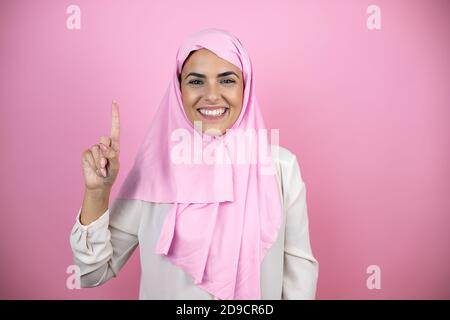 Giovane bella donna araba che indossa hijab islamico su rosa isolato sfondo che mostra e punta verso l'alto con le dita numero uno mentre sorridente sicuro Foto Stock