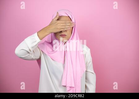 Giovane bella donna araba che indossa hijab islamico su rosa isolato sfondo serio e coprendo i suoi occhi con la mano Foto Stock