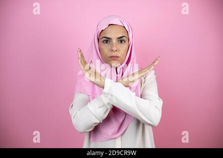 Giovane bella donna araba che indossa hijab islamico su sfondo rosa isolato espressione di rifiuto che attraversa le armi facendo segno negativo, faccia arrabbiata Foto Stock