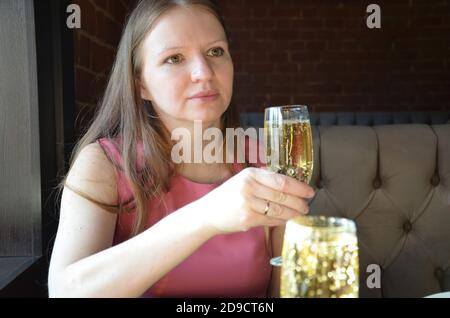 bella ragazza bionda che tiene un bicchiere di champagne o vino, bere champagne in un ristorante, in un abito rosa sera, in una data o riunione Foto Stock