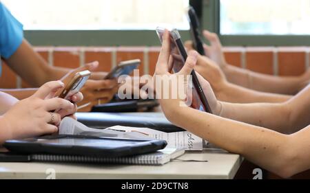 Un tavolo di studenti delle scuole superiori che tengono tutti più dispositivi digitali, come telefoni e tablet, durante una lezione di classe. Istruzione contemporanea e conn Foto Stock