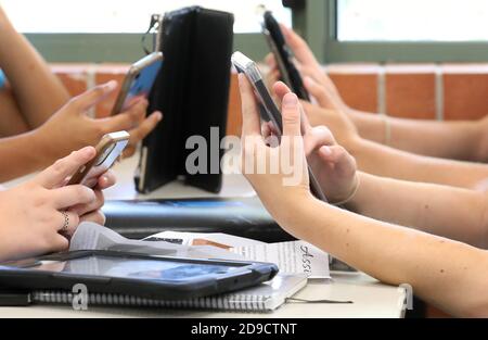 Studenti che tengono e utilizzano una gamma di dispositivi digitali. I tablet e la tecnologia dei telefoni cellulari sono gestiti dagli studenti in una scrivania in un'istruzione scolastica Foto Stock