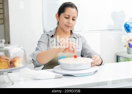 Donna ispanica nella sua pasticceria che decora una torta-donna che mette ciliegia sulla torta con panna montata - donna intraprendente Foto Stock